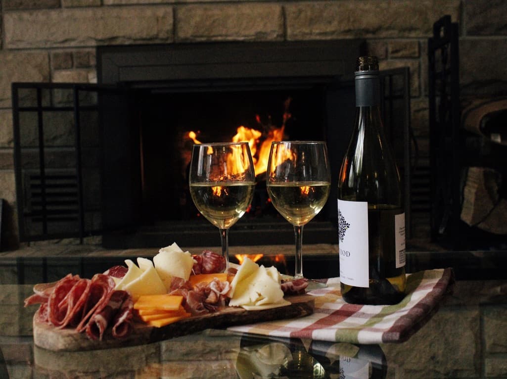 A glass of wine and a cheese platter on a table beside a glowing fireplace